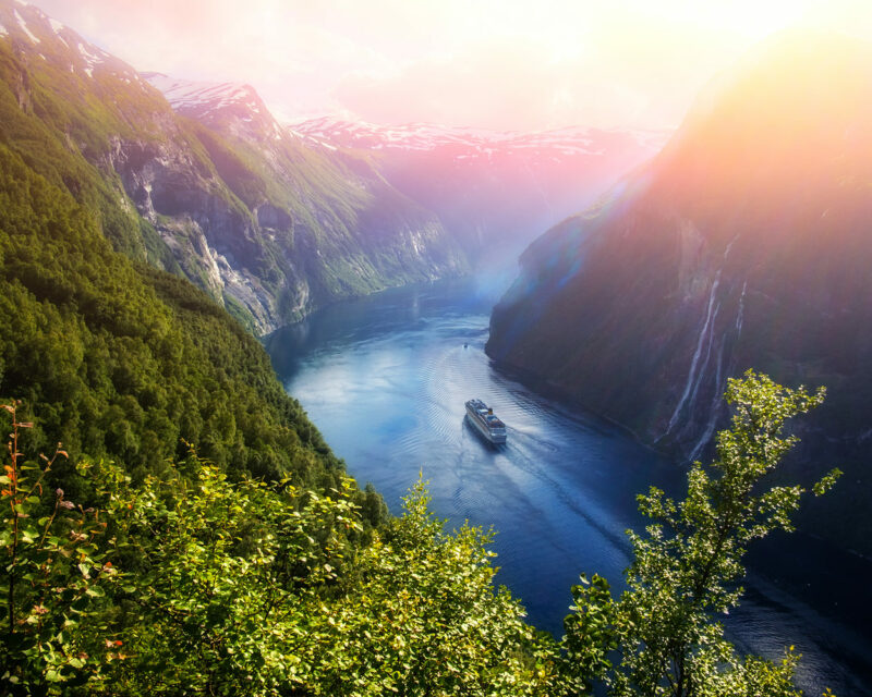 A river with a ship sailing into the sunset surround by lush vegetation.